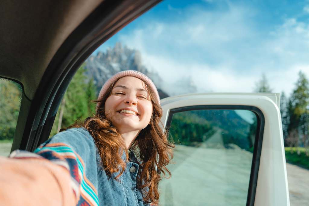 Road Trip Tips: Protecting Your Belongings While Traveling for Assignments A woman smiling and taking a selfie in a car, with a mountainous landscape visible through the window. The article provides various tips for healthcare travelers to protect their belongings during road trips for assignments