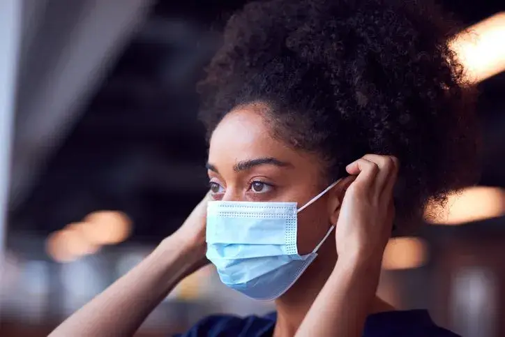 What To Do If You’ve Been Exposed to COVID-19 Female Doctor In Scrubs Putting On Face Mask Under Pressure In Busy Hospital During Health Pandemic