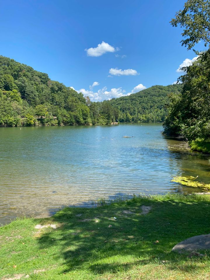 Must-See Places in Kentucky on Your Travel Assignment Fishpond Lake near Jenkins, Kentucky. The surrounding area features green, tree-covered hillsides and a clear blue sky with some clouds. The webpage text is a blog post titled "Must-See Places in Kentucky on Your Travel Assignment", which discusses various natural attractions in the state, including Mammoth Cave National Park, Cumberland Falls State Resort Park, and Red River Gorge.