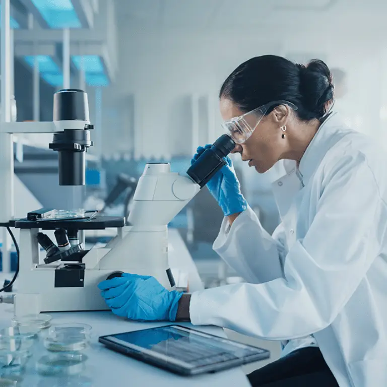 Life sciences professional wearing protective equipment examining a sample through a microscope in a laboratory.