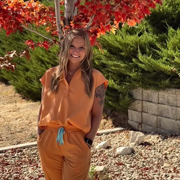 Healthcare professional in orange scrubs smiling outdoors beneath colorful autumn leaves.