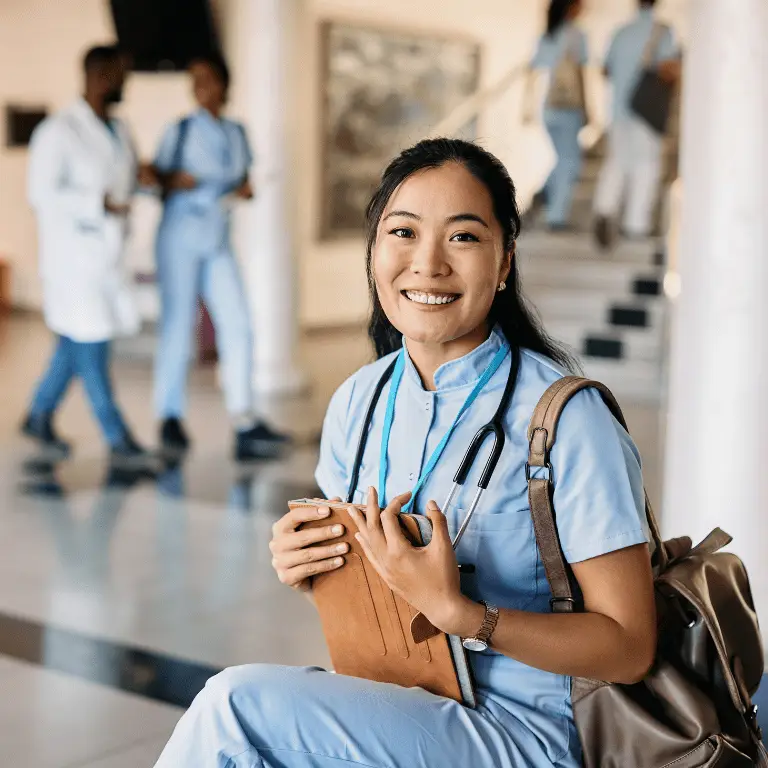 Healthcare professional in blue scrubs with a stethoscope and backpack smiling in a healthcare facility.