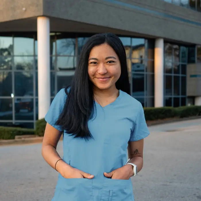 Healthcare professional in blue scrubs smiling outside a healthcare facility.