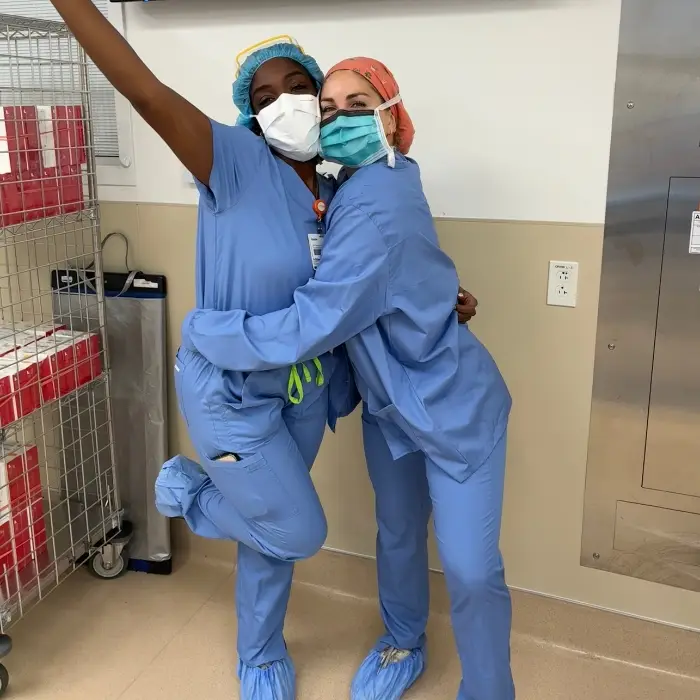 Two operating room nurses in blue scrubs, surgical caps, and face masks hugging in a hospital.