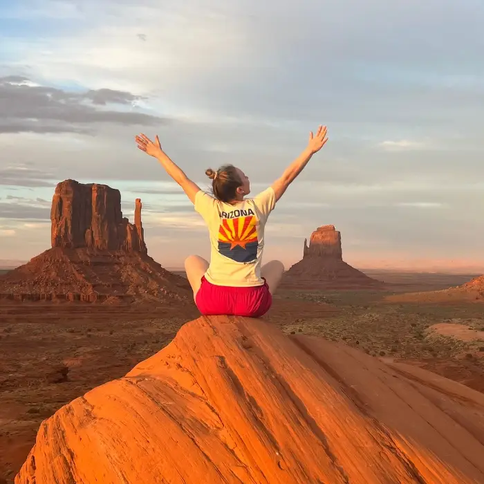 TotalMed healthcare traveler sitting on a red rock overlook with arms raised above the desert landscape of Monument Valley.